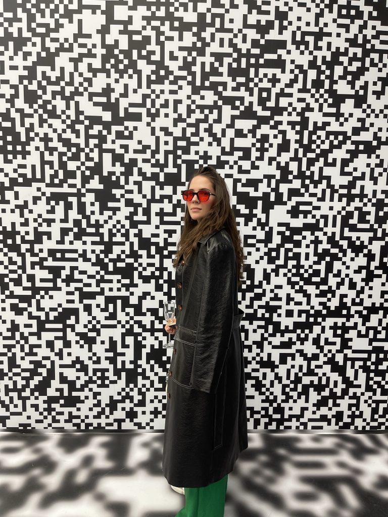 our new co-author pauline, a brunette girl, is standing in front of a black and white pixelated wall. The photo was taken at an event for web3 fashion brands